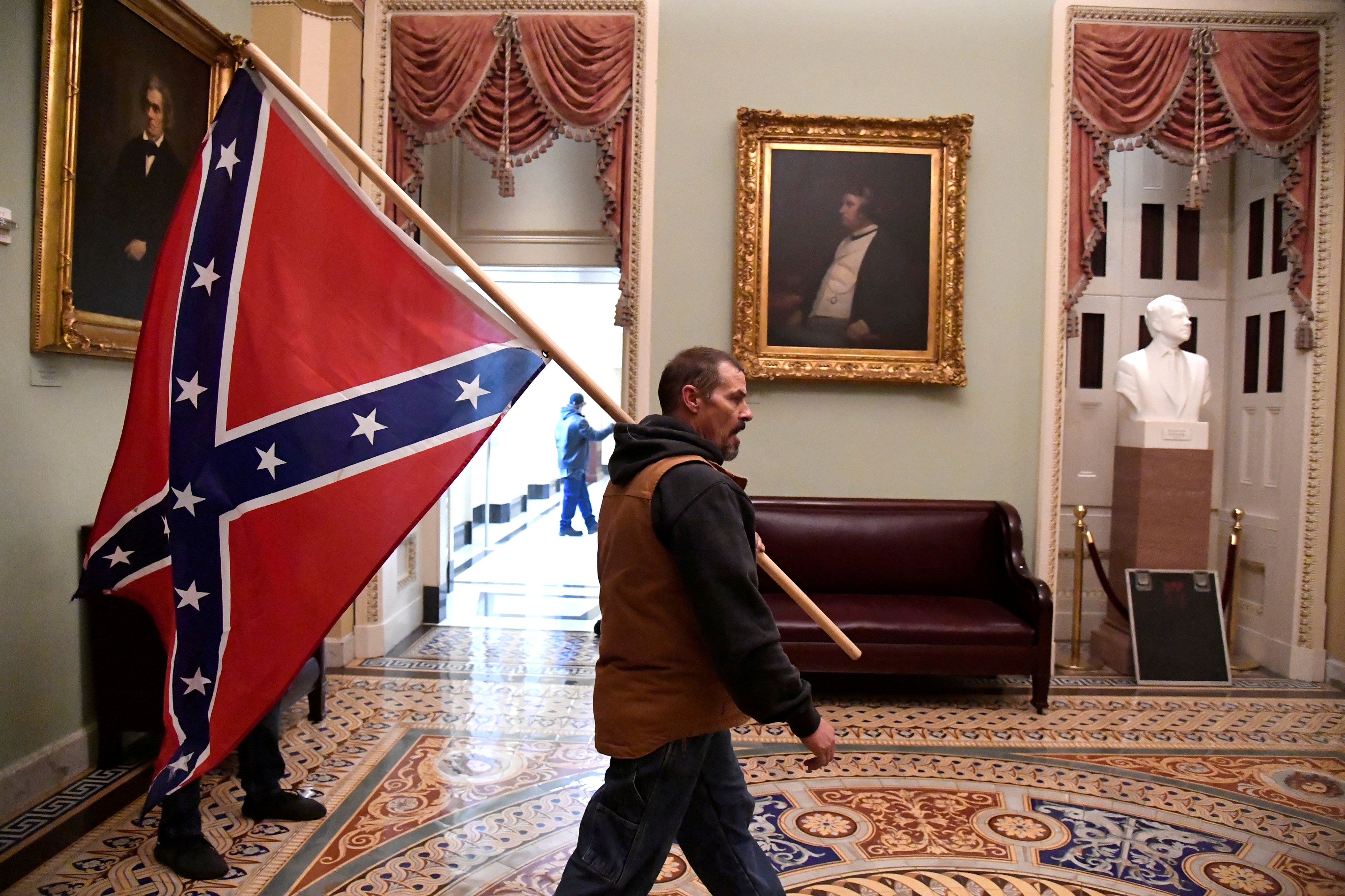 A profile photograph of a white man. He is walking through the United States Capitol on the day of the Jan 6 Insurrection carrying a large Confederate flag.